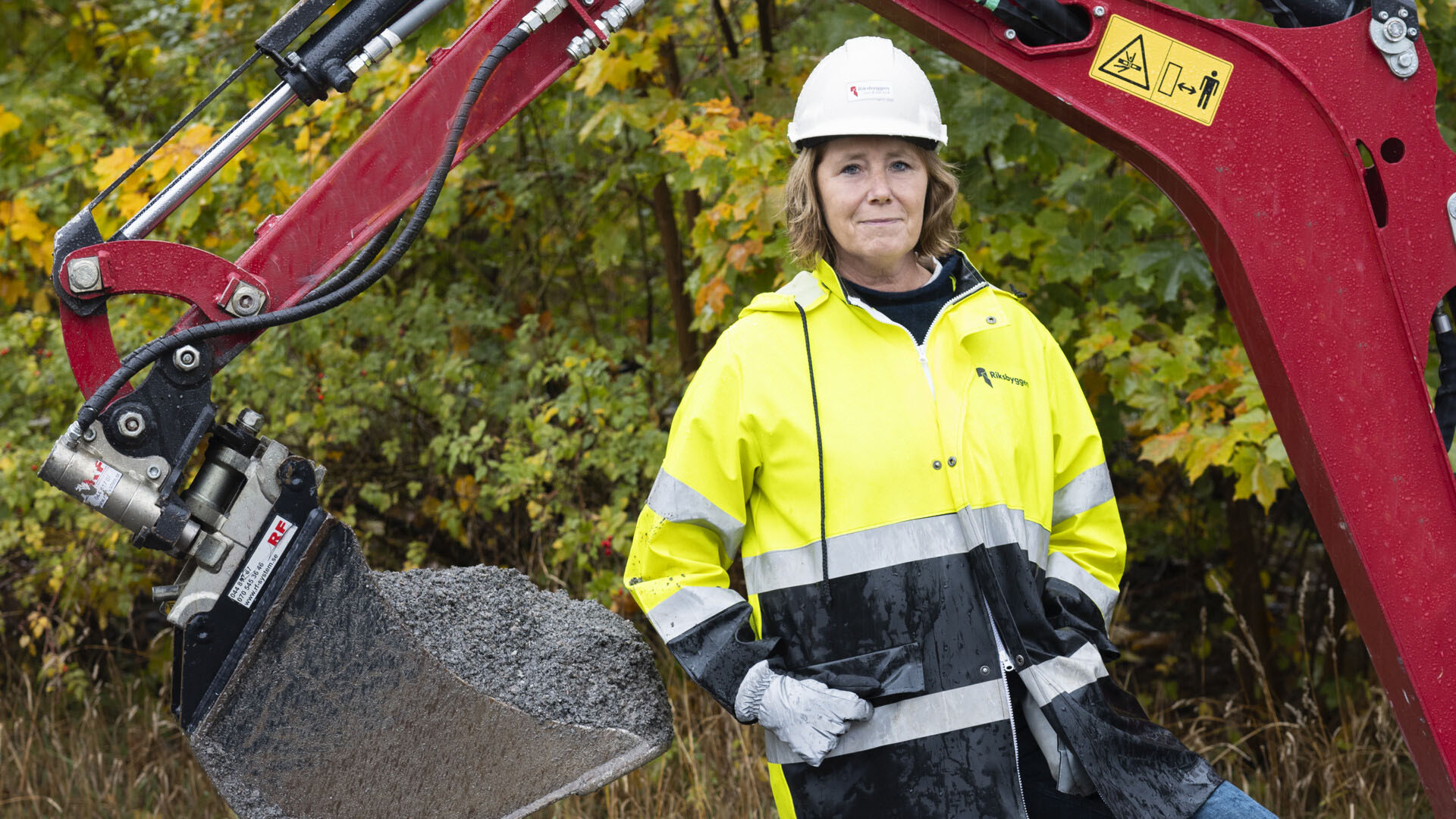 Riksbyggens vd Johanna Frelin iklädd en gul arbetsjacka stående framför en röd grävskopa på en byggplats